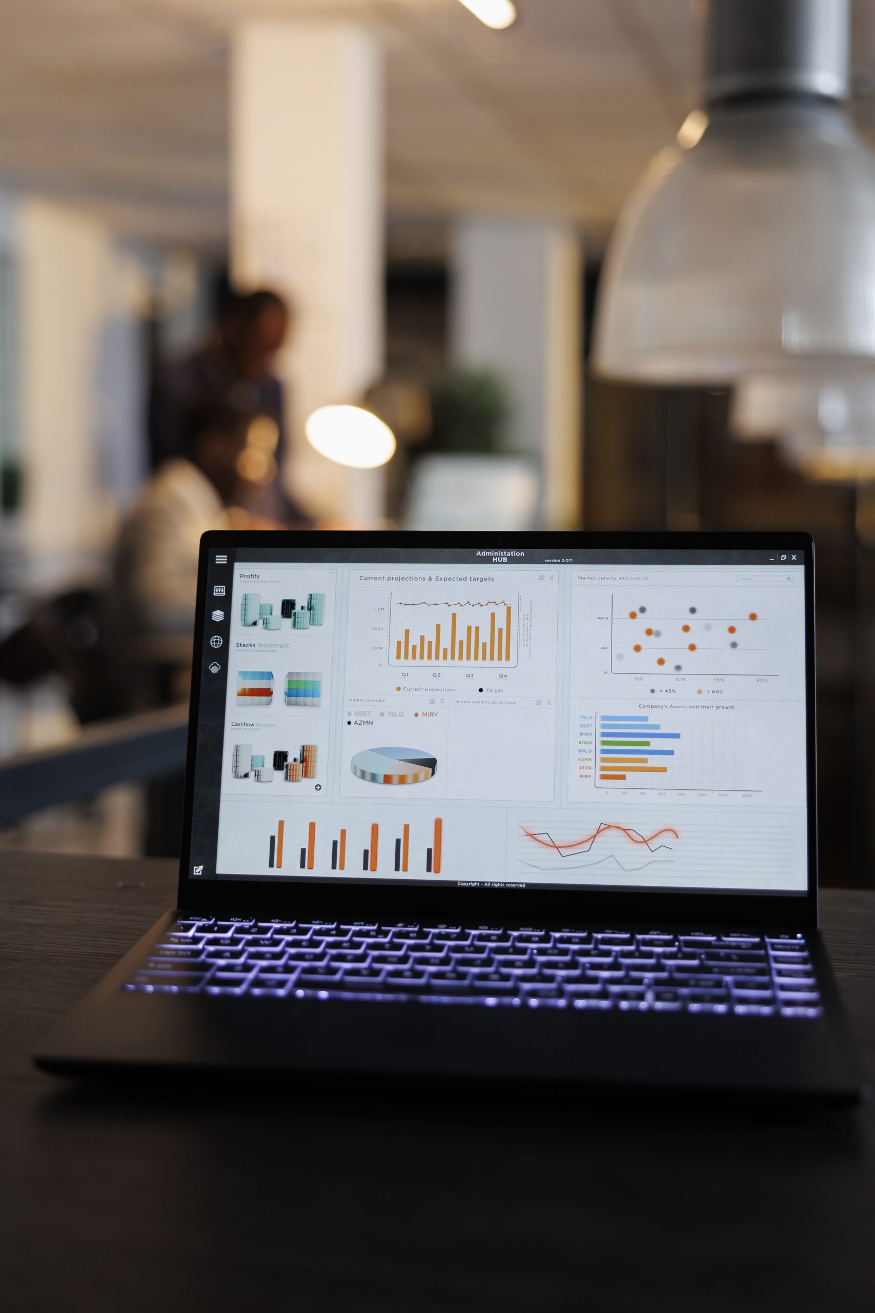 Laptop computer with marketing statistics graphs on screen, standing on table in startup office, coworkers discussing company strategy in background. Employees working overtime in startup office